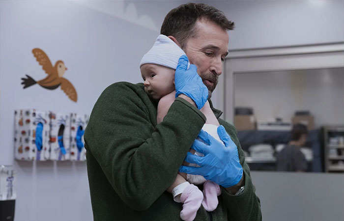Man wearing gloves holding a newborn baby indoors, related to The Pitt season 2 episode 2 preview and release details