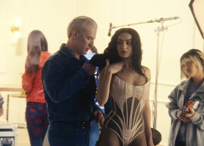 Charli XCX backstage wearing a racy corset outfit being styled during a braless photo shoot session.