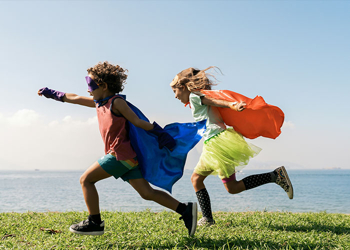 Two children running by the water wearing colorful superhero capes, symbolizing nannies and the internet raising kids.