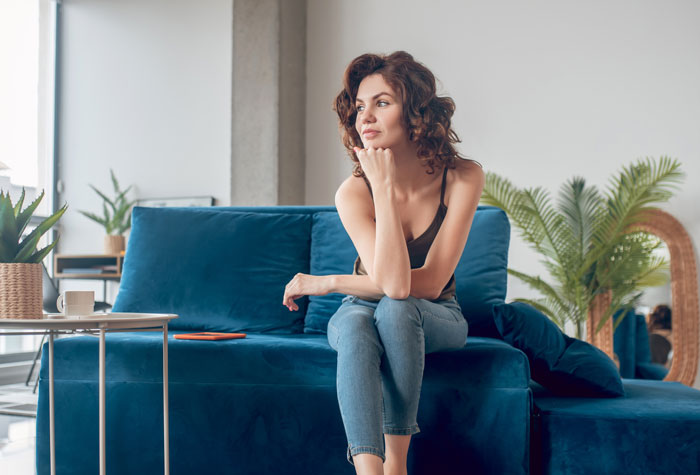 Woman sitting on a blue couch, looking thoughtful and holding her chin, with a phone on the table nearby.