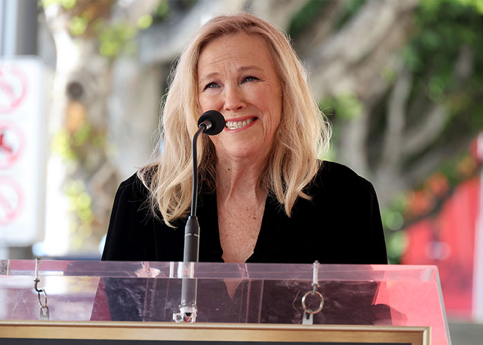 Catherine O'Hara speaking at a podium outdoors, smiling warmly with trees and blurred background behind her. Catherine O'Hara speaking at a podium outdoors, smiling warmly with trees and blurred background behind her.