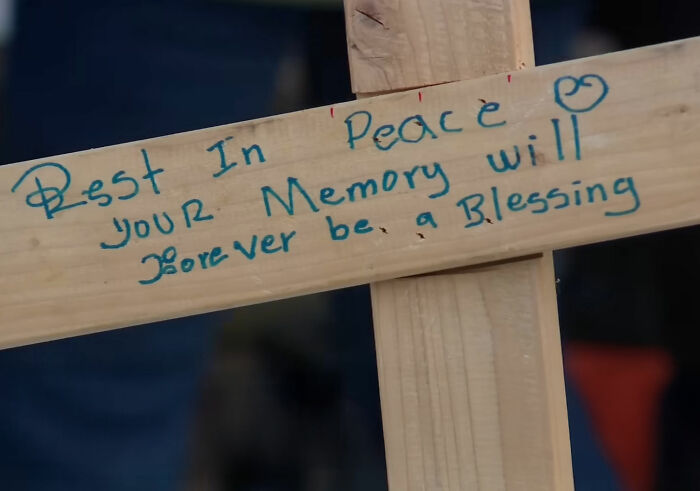 Wooden memorial cross with handwritten message in blue ink honoring a mom slain by ICE agent, family expresses grief and anger.