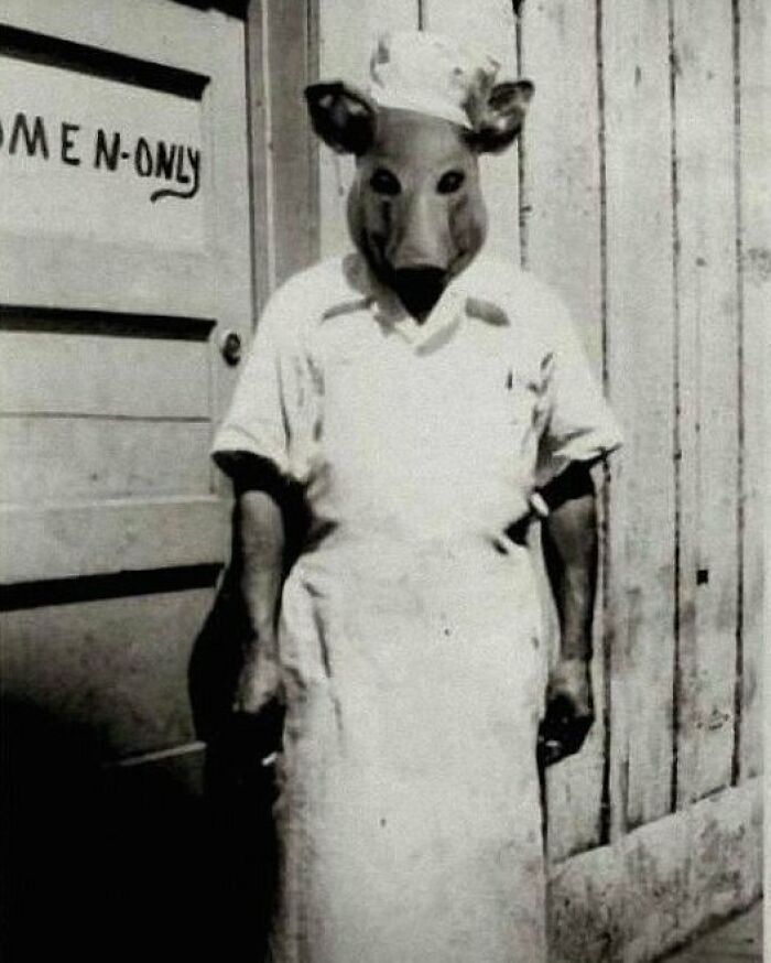 Black and white vintage photo of a person wearing a creepy animal mask and chef uniform standing by a women-only door.