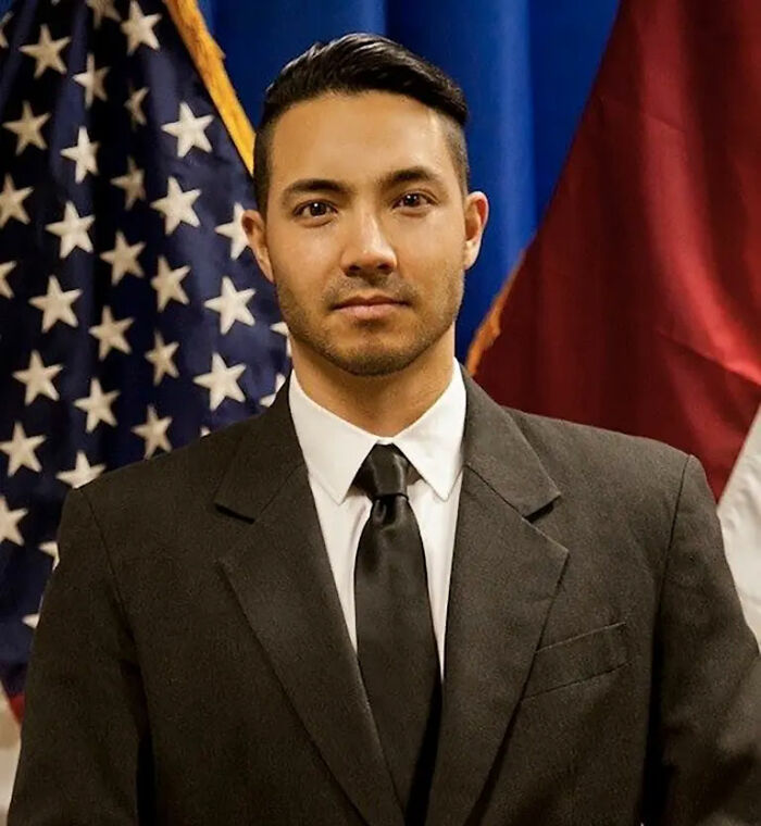 Marine veteran in formal suit standing in front of American and military flags, reflecting a somber and respectful moment.