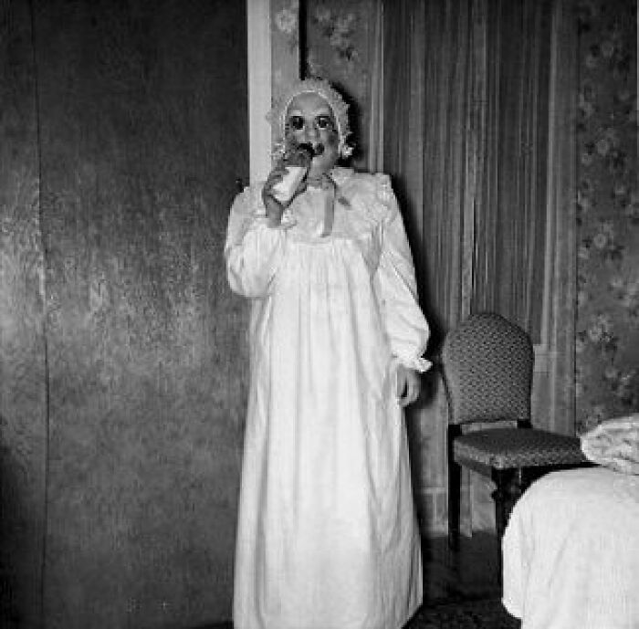Old black and white creepy photo of a person in a vintage nightgown and bonnet holding a bottle in a dimly lit room.