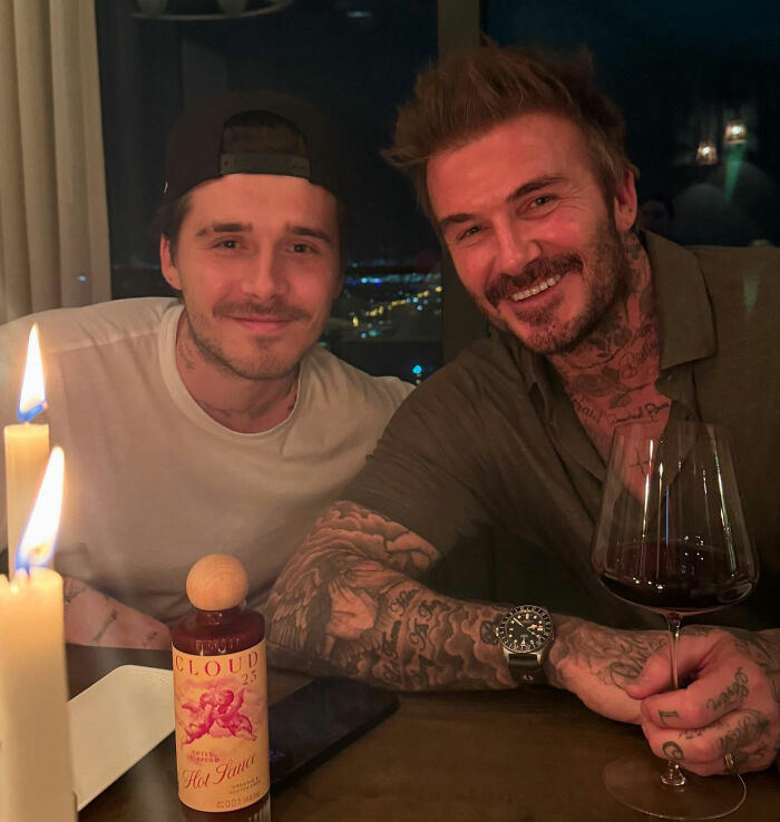 Brooklyn Beckham and David Beckham smiling at a dinner table with wine and candles amid family feud talk.