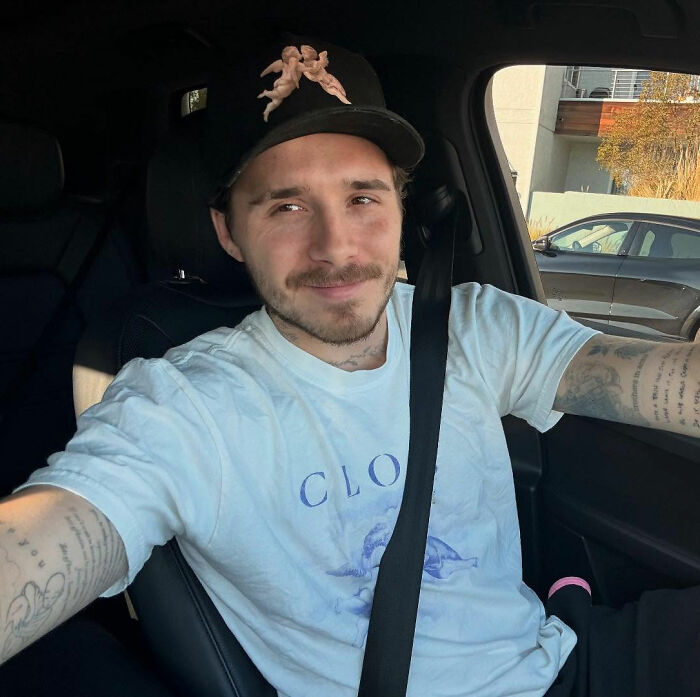 Brooklyn Beckham wearing a cap and white shirt, sitting in a car while taking a selfie amid family feud legal dispute.