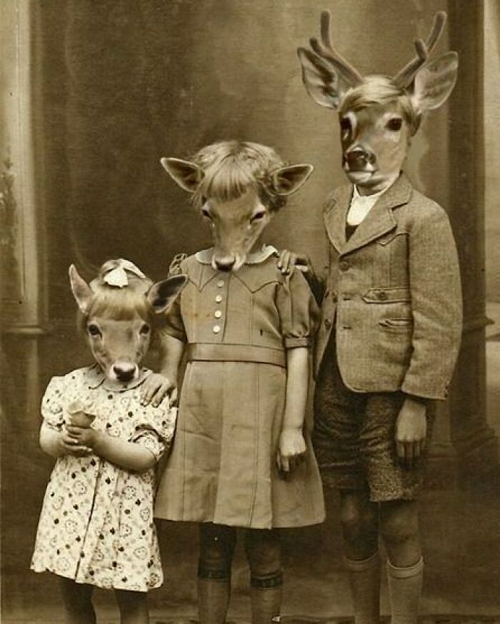 Vintage black and white photo of three children with deer heads in old-fashioned clothing, odd and creepy style.