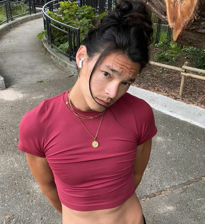 Young man with dark hair in a bun wearing a red cropped shirt, outdoors near a curved path and plants in San Francisco.
