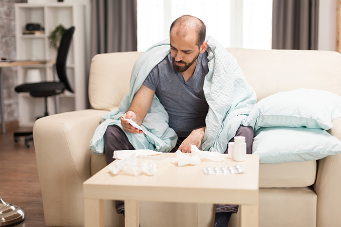Man wrapped in blanket on sofa, looking at thermometer and medicine, depicting cancer suffering and illness symptoms.