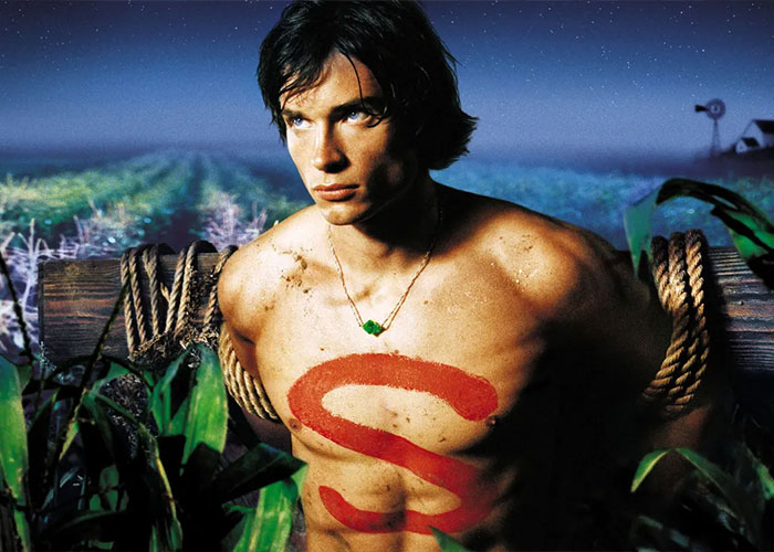 Young 2000s teen heartthrob with shirtless torso and red S painted, tied to wooden post in a cornfield at night.