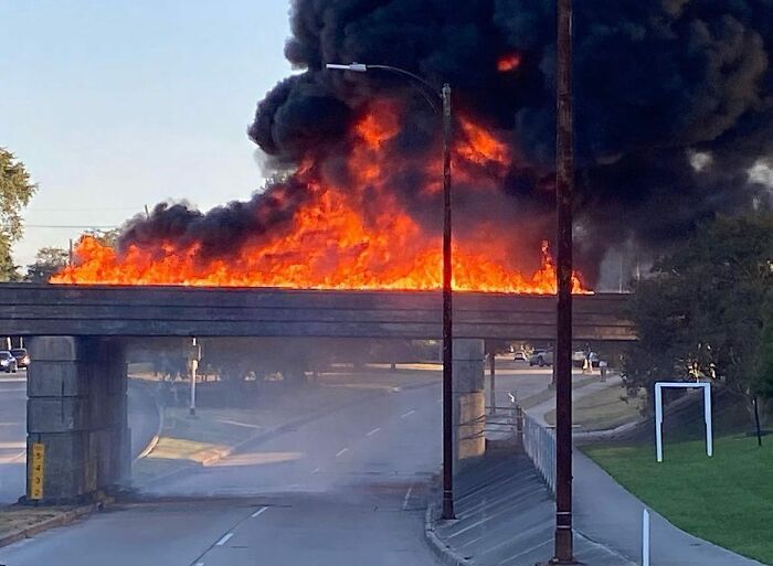 Bridge on fire with thick black smoke rising above a city street, showing a weird urban street scene found by people.