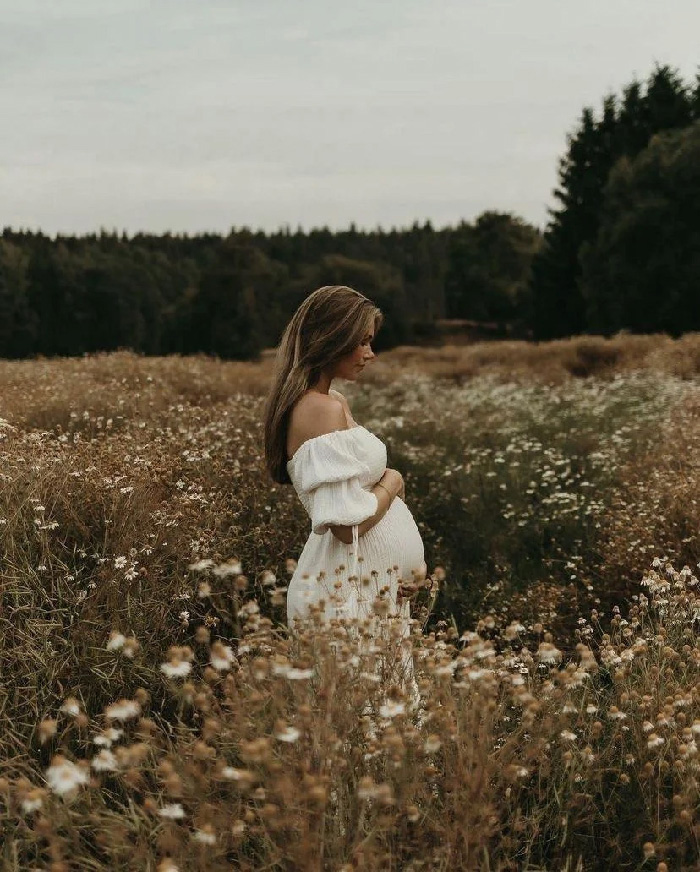 Pregnant woman in white off-shoulder dress cradling belly in wildflower field, maternity photoshoot, soft golden light.