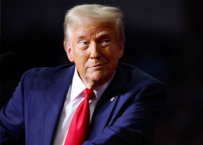 Portrait of a man in a dark suit and red tie, looking to the side with a thoughtful expression in a dim setting.