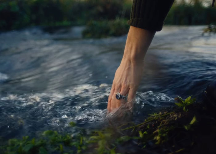 Hand with sapphire ring touching flowing water by a small stream, evoking calm and nature in soft daylight.