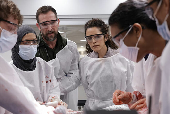 Medical team in protective gear examining a body, highlighting The Pitt Season 2 Episode 1 recap key moments.