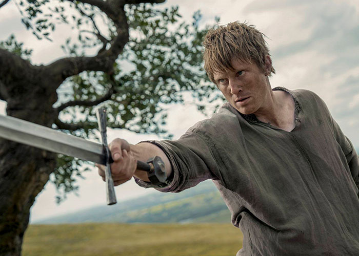Young man in medieval clothing holding a sword outdoors, related to George RR Martin and A Knight of the Seven Kingdoms.