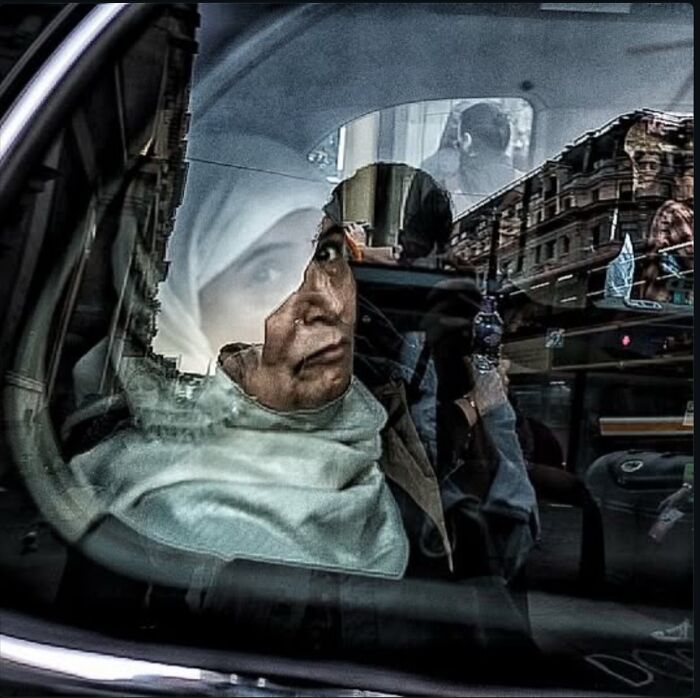 Candid street photo capturing a reflective moment of a woman wearing a headscarf inside a vehicle on a city street.