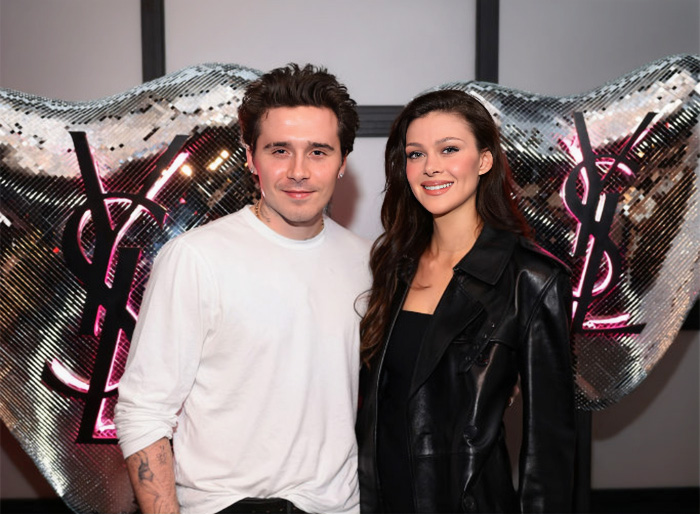Brooklyn Beckham and Nicola Peltz posing together at a stylish event with yin-yang logo decorations behind them.