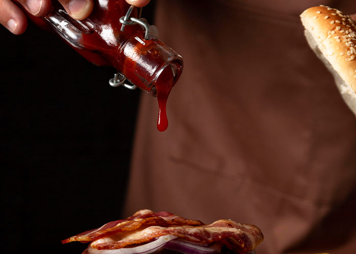 Hand pouring sauce from a glass bottle onto bacon and onions in a sandwich, illustrating revenge acts so perfect.