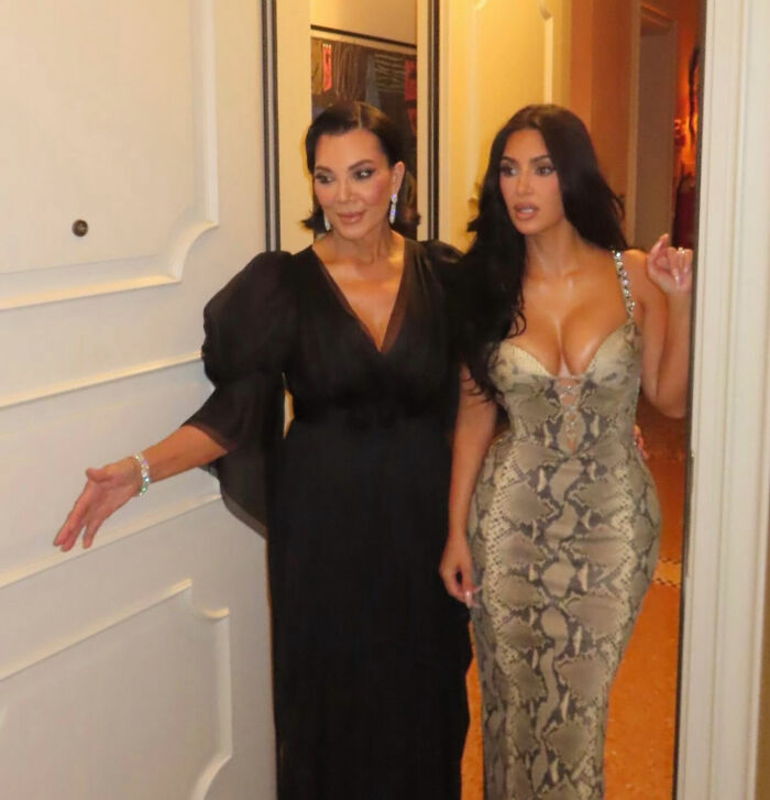 Kim Kardashian in a snakeskin dress with another woman in a black dress, pictured in an elegant indoor setting.