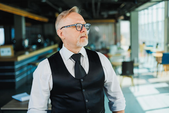 Older male service industry worker wearing glasses and a vest, experiencing an awkward moment in a bright restaurant setting
