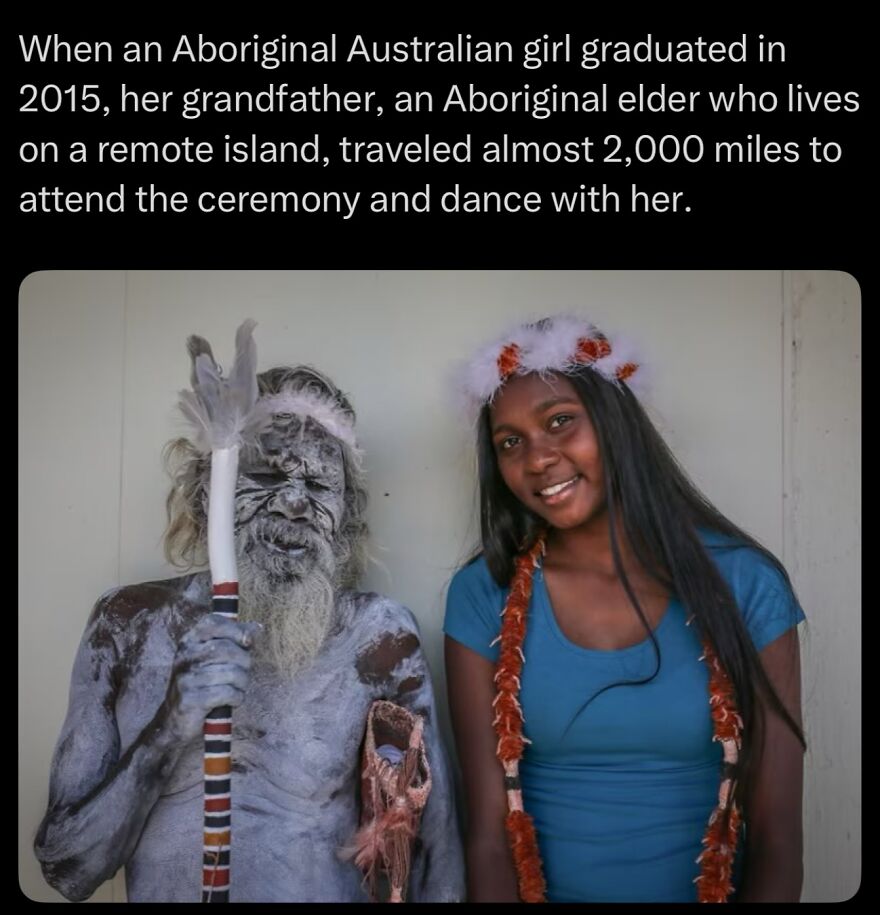 Aboriginal elder covered in body paint holding a staff next to a smiling Aboriginal girl wearing a flower headband and necklace