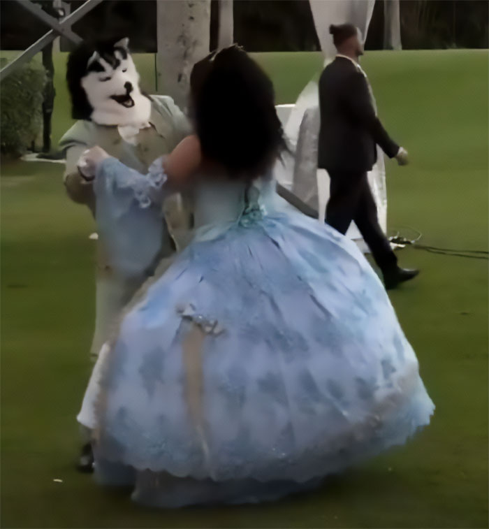 Person in dog mask dancing with woman in old-time blue costume at a creepy Mar-a-Lago event outdoors on grass.
