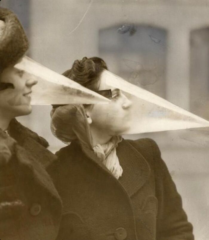Two women in old black and white photo wearing long, pointed odd and creepy face masks outdoors.