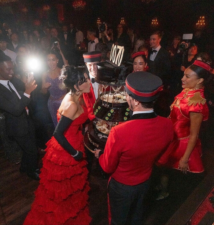 Kim Kardashian at a party with people dressed in red, photographers capturing the moment amid a large cake celebration.