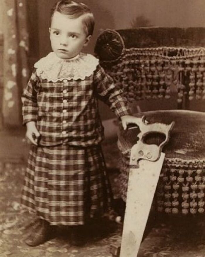 Vintage black and white creepy photo of a young child in old-fashioned clothing holding a large saw odd black and white pic.