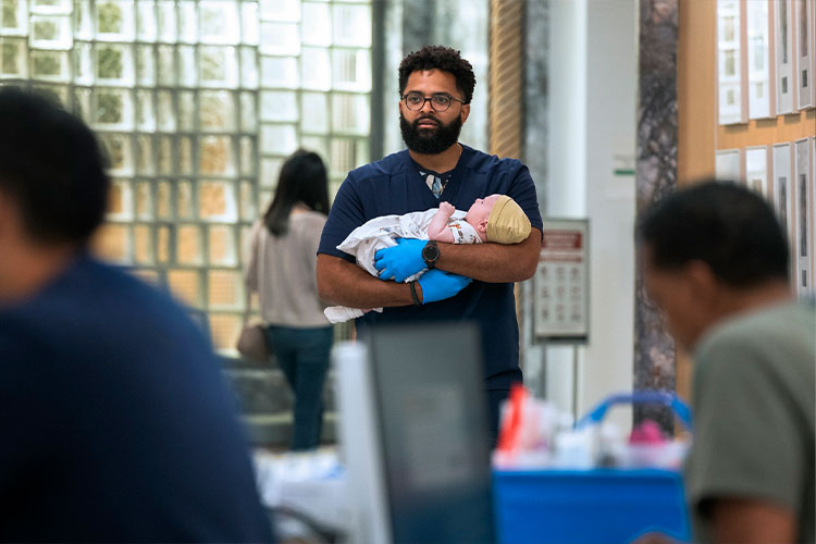 Man in medical scrubs holding a newborn baby indoors, illustrating The Pitt season 2 episode 1 recap scene.