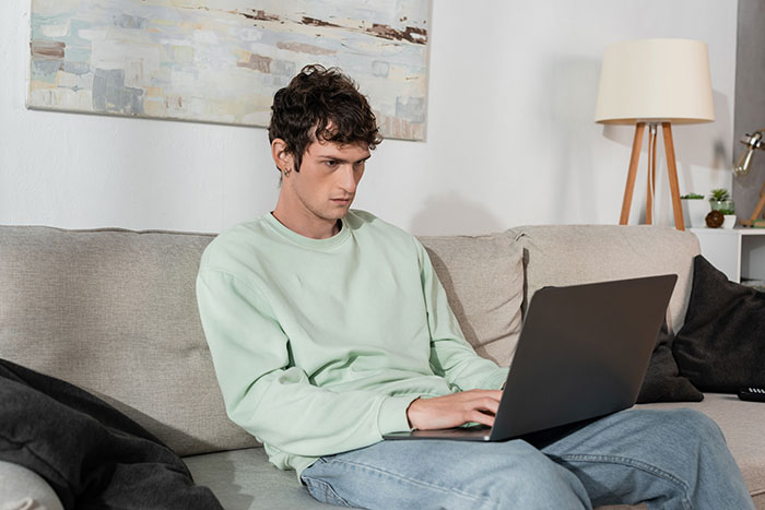 Young man sitting on couch using laptop, reflecting on challenges after dad gets accused of horrific things by his own kids