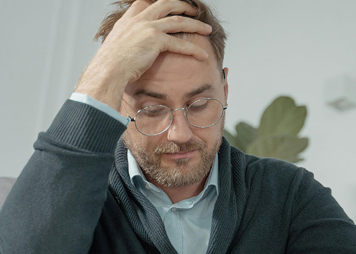Man with glasses looking down and holding head, reflecting on life changes after discovering not biological parent.