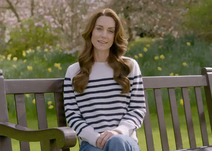 Kate Middleton sitting on a park bench wearing a striped sweater, surrounded by green grass and flowers in spring.