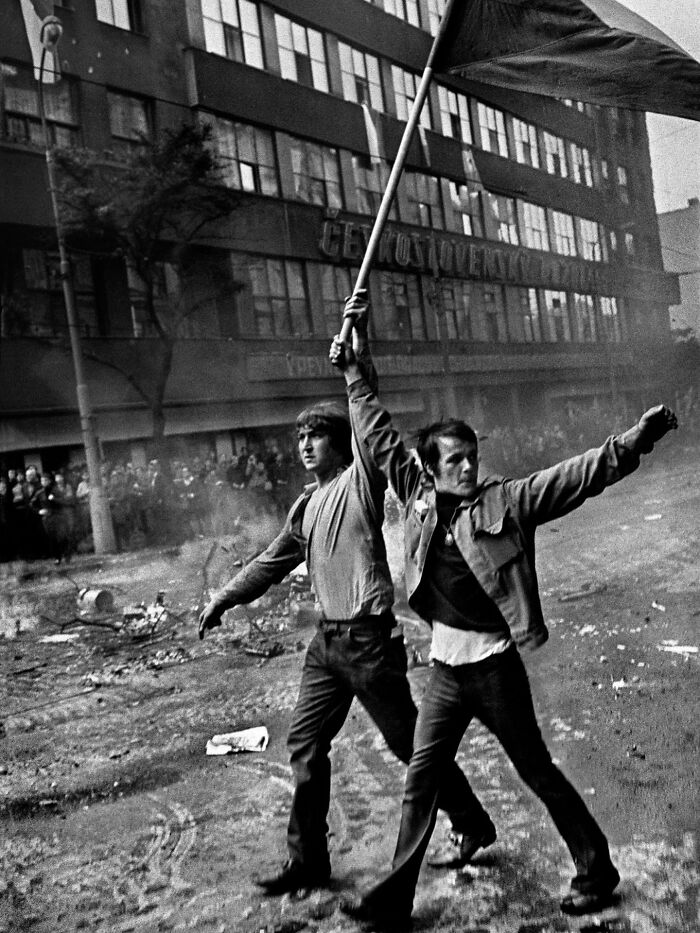 Two men in a street photo raising a flag amid chaos, capturing life’s unexpected and beautiful moments in a black and white scene.