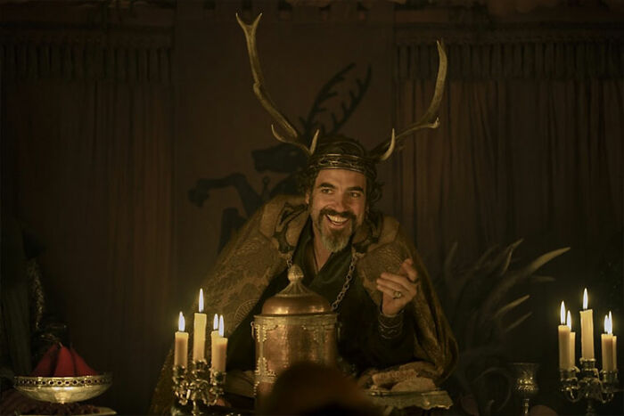 A Knight Of The Seven Kingdoms cast member smiling in medieval attire with candles and a crown in a dimly lit room. A Knight Of The Seven Kingdoms cast member smiling in medieval attire with candles and a crown in a dimly lit room.