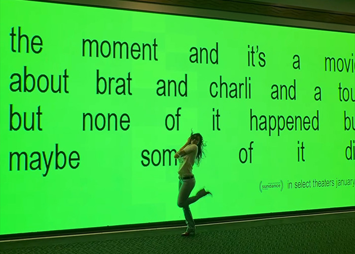 Person posing in front of a large bright green screen with text, evoking Charli XCX racy braless photo dump vibe.