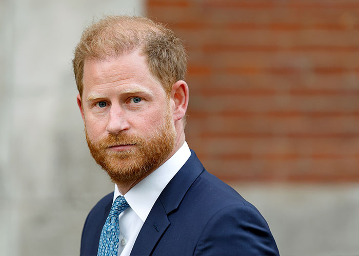 Prince Harry in a navy suit with a blue patterned tie appearing serious against a blurred brick background during trial.