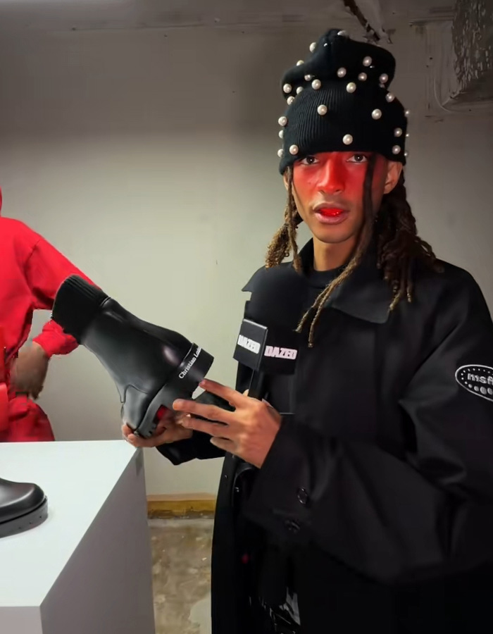 Person wearing a pearl-studded black hat holding a Christian Louboutin boot during a fashion event in an indoor setting. Person wearing a pearl-studded black hat holding a Christian Louboutin boot during a fashion event in an indoor setting.