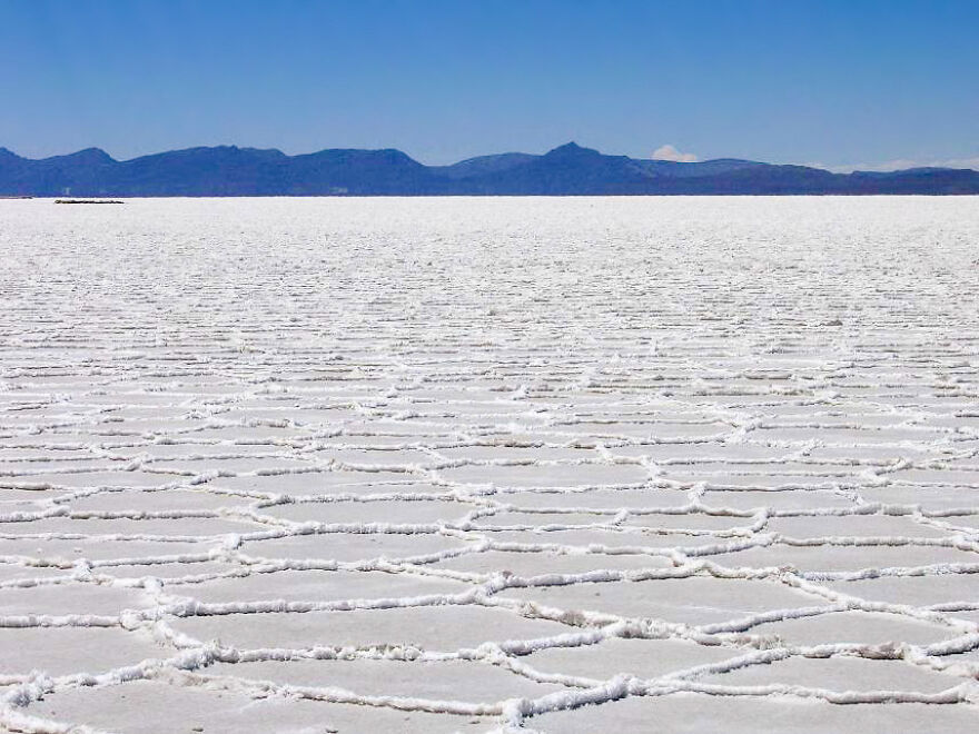 Vast salt flats with hexagonal patterns under clear blue sky and mountains, a must-see place to visit once in life.