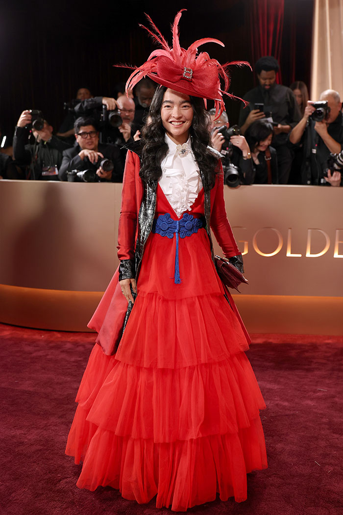 Woman in a layered red gown with feathered hat on the red carpet at the 2026 Golden Globes fashion disasters event.