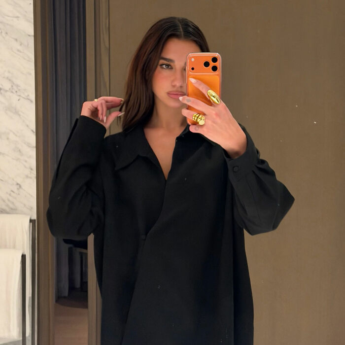 Dua Lipa taking a mirror selfie wearing a black outfit, showing gold rings and holding an orange phone.