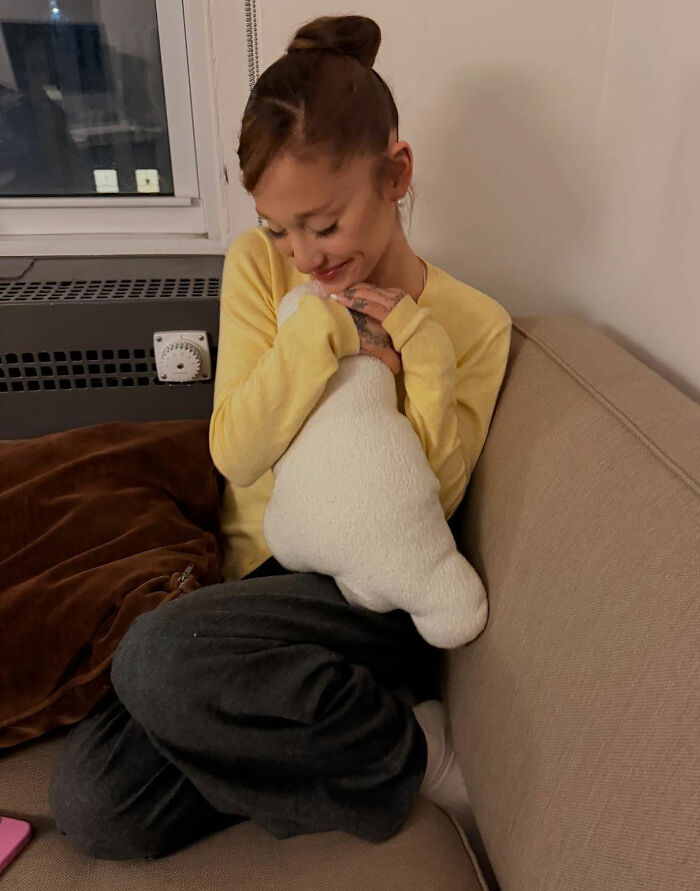 Ariana Grande sitting on a couch in casual clothes, smiling and hugging a white plush toy indoors. Ariana Grande sitting on a couch in casual clothes, smiling and hugging a white plush toy indoors.