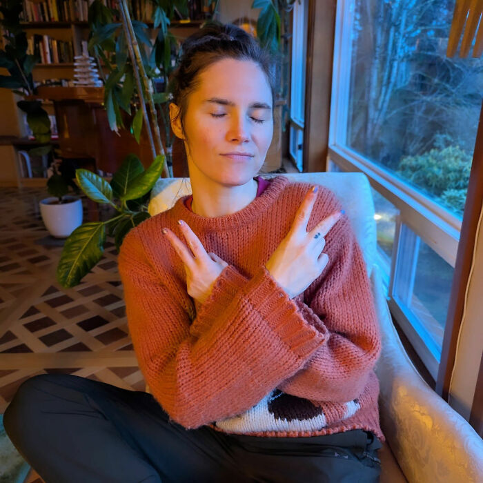 Amanda Knox indoors wearing a rust-colored sweater, sitting with eyes closed and hands crossed, relating to cancel culture feud.