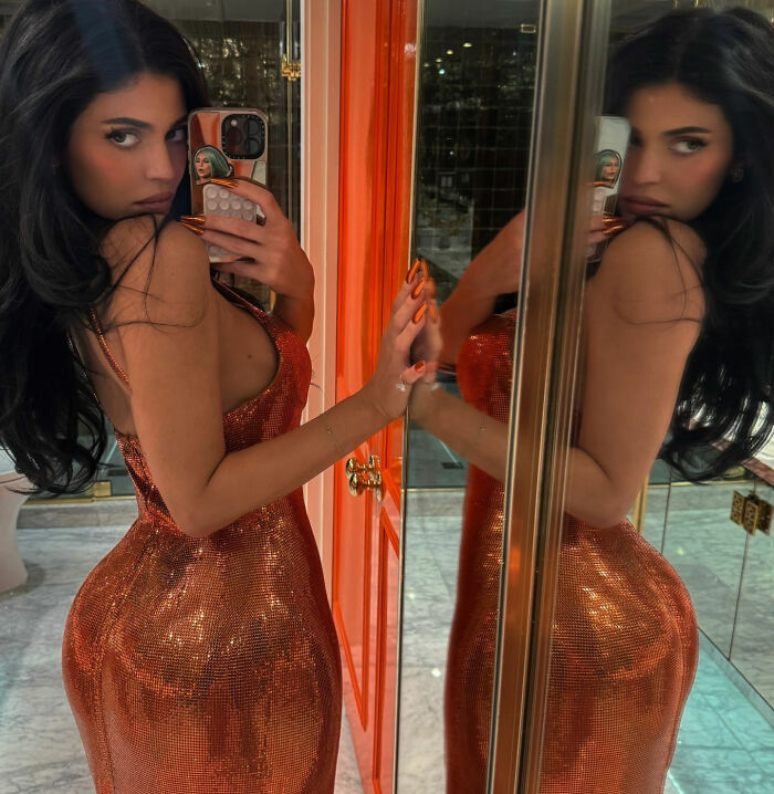 Woman in a sparkling orange dress taking a mirror selfie, highlighting Kylie&rsquo;s BBL in a stylish indoor setting.