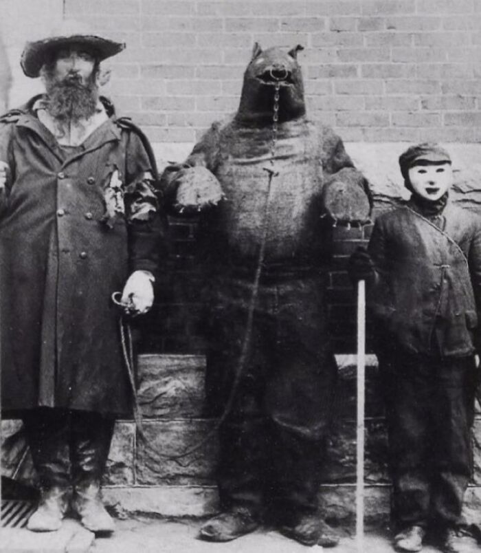 Three people in old black and white photo wearing odd and creepy costumes standing against a brick wall.