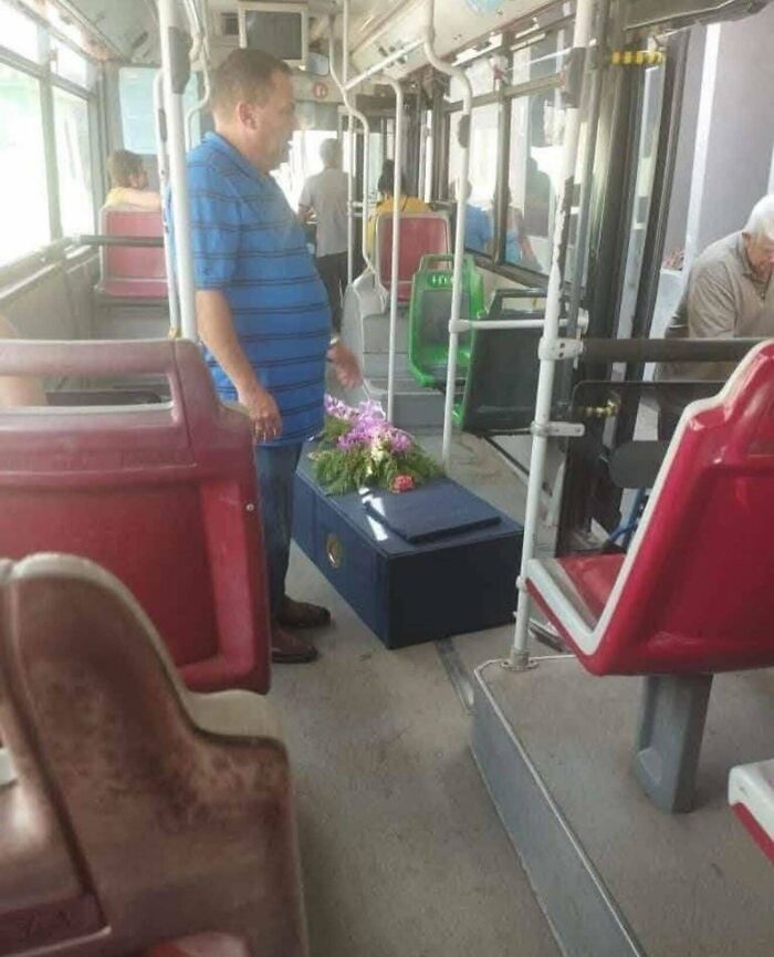 Man standing next to a blue coffin with flowers inside a public bus, an interesting but creepy photo scene.