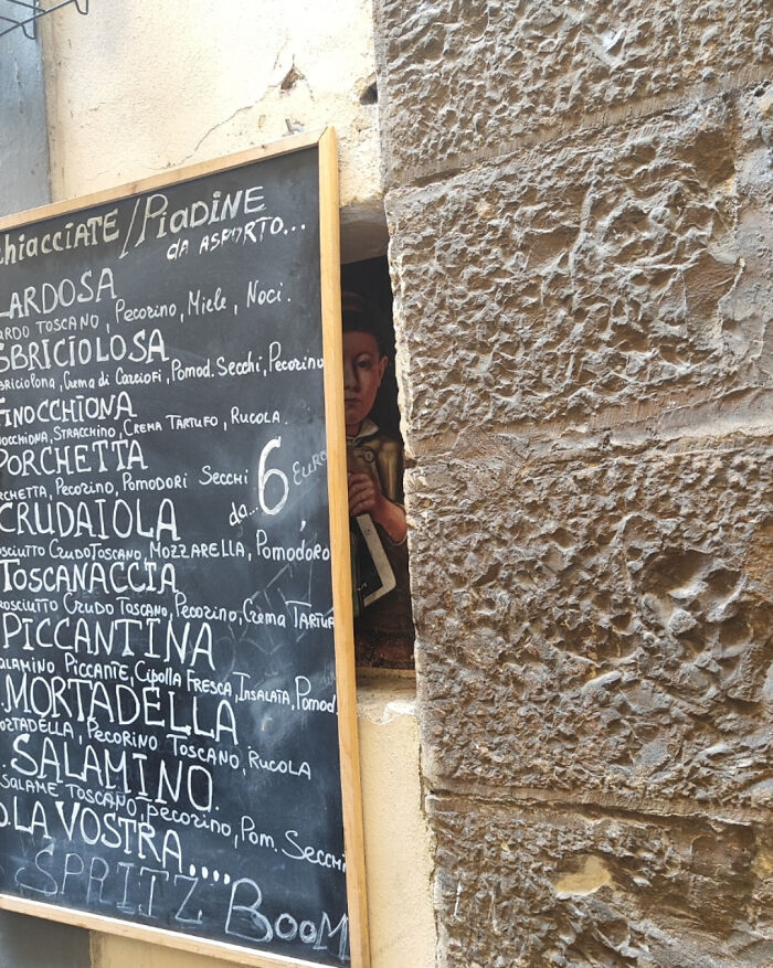 Chalkboard menu with Italian food names beside a stone wall and a partially hidden creepy painted face peeking through a small opening.
