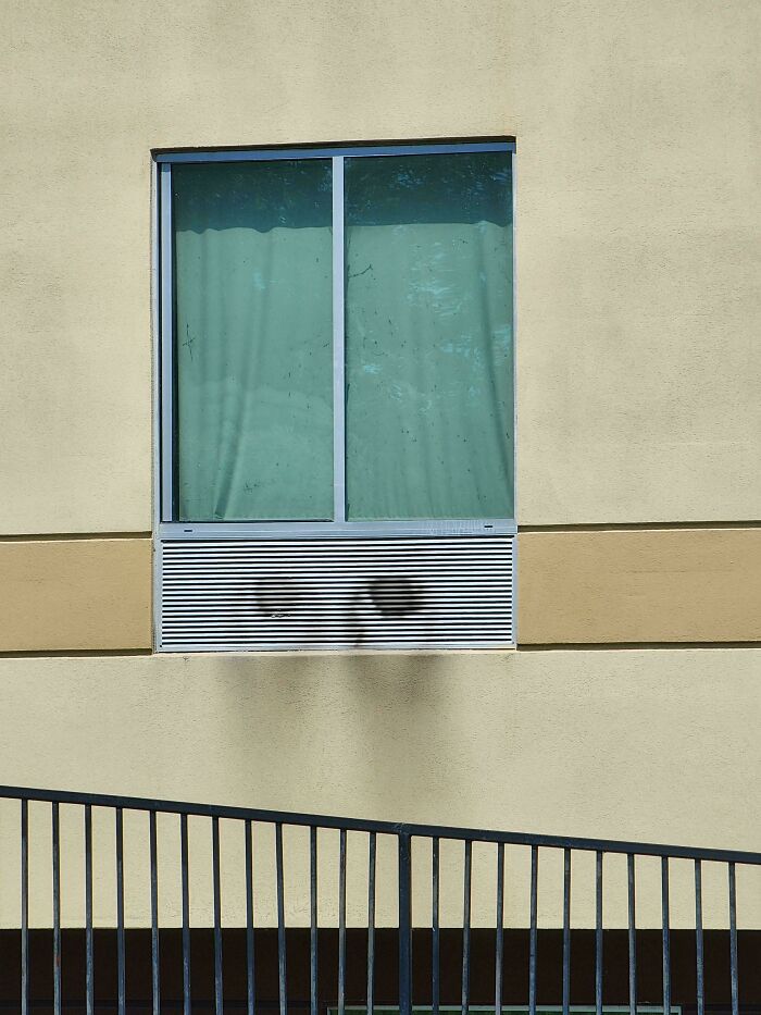 Window air vent casting interesting but creepy shadow resembling two dark eyes on a beige wall outdoors.